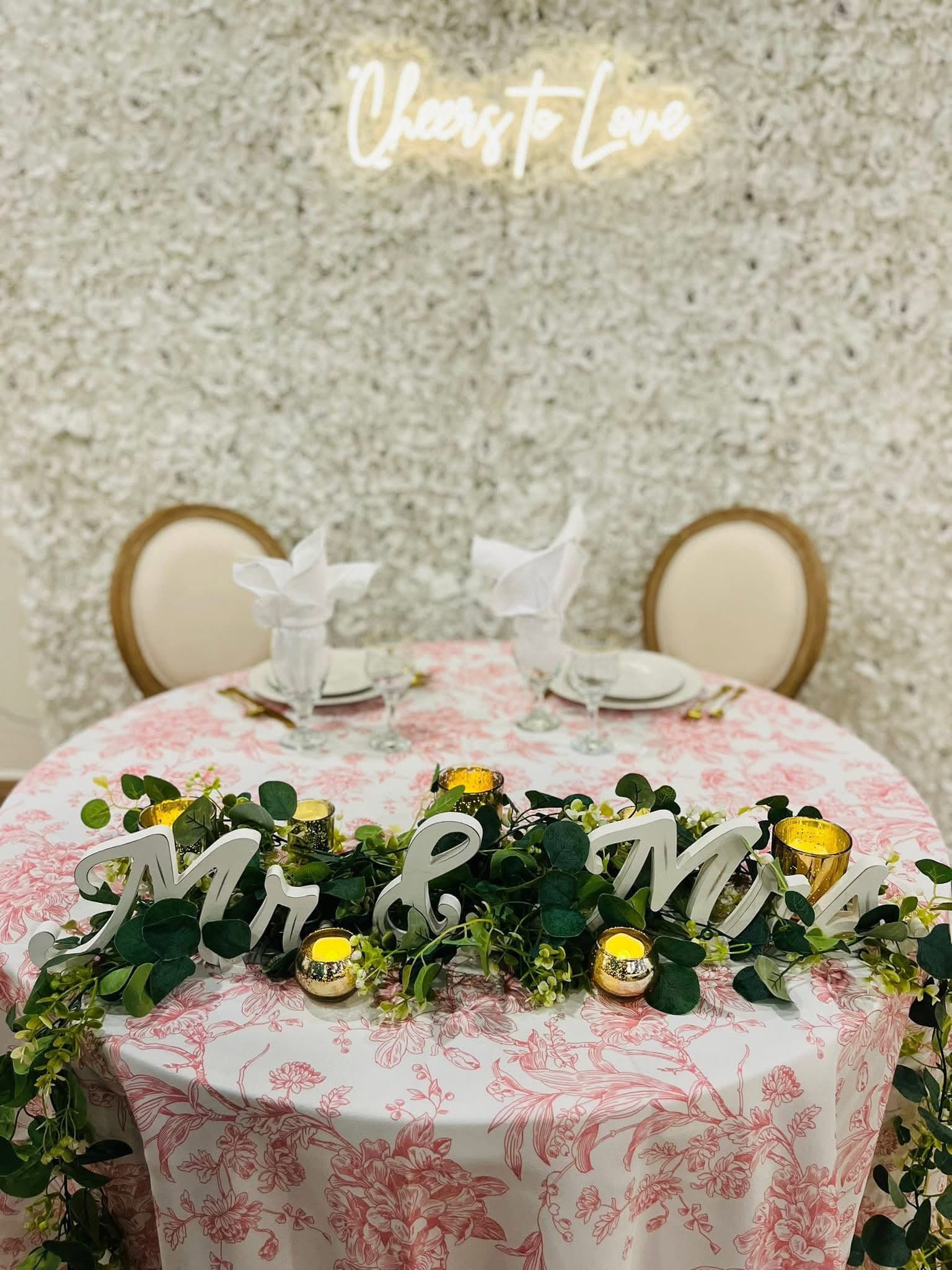 Sweetheart table with a Cheers to Love neon sign and floral wall backdrop at Becker Hill